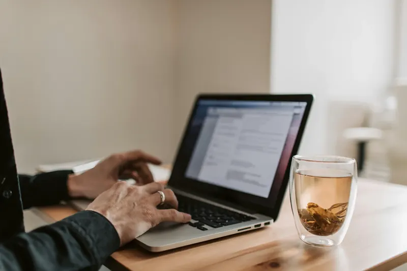 Hände tippen auf einem Laptop, daneben ein Glas Tee